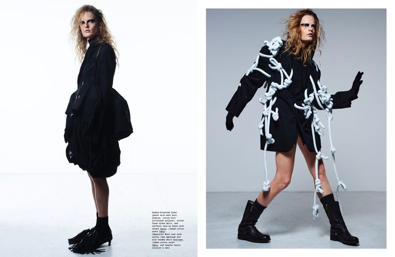 Editorial fashion shot: Woman in black jacket with decorative white rope details and black boots poses in a bright photo studio.