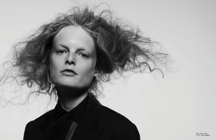 Black and white close-up portrait of a young woman with tousled hair, wearing a dark blouse against a bright white background.