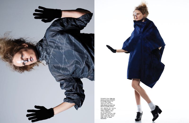Two fashion models pose in a studio wearing avant-garde, structural clothing; one in dark blue and another in a dark grey with patterns.