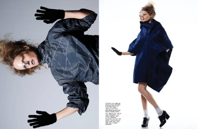 Two fashion models pose in a studio wearing avant-garde, structural clothing; one in dark blue and another in a dark grey with patterns.