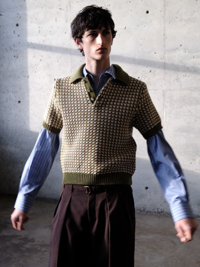 Male fashion model poses in front of a concrete wall, wearing a short sleeve polo sweater, collared shirt, and brown slacks with a belt.