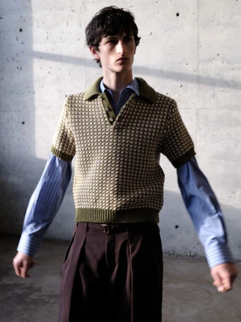 Male fashion model poses in front of a concrete wall, wearing a short sleeve polo sweater, collared shirt, and brown slacks with a belt.