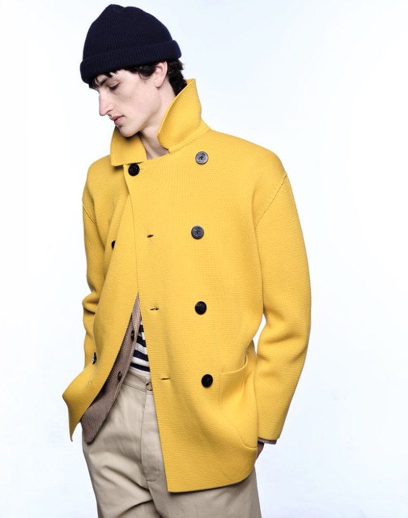 Man in yellow pea coat and navy beanie in a studio shot. Neutral background. He is looking down and away from the camera.