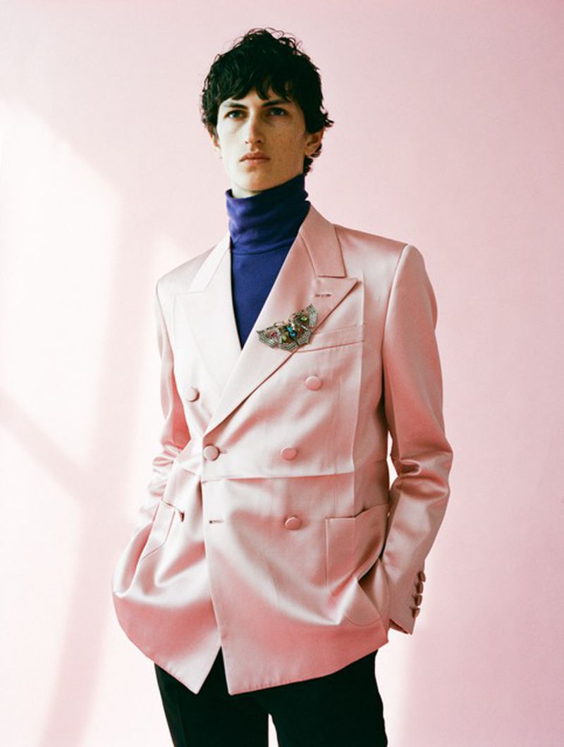Fashion portrait of a young male adult wearing a pink satin blazer, dark blue turtleneck, and dark pants against a pink background.