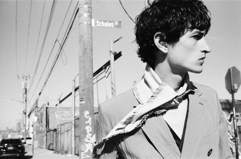 Portrait of a young adult male in an outdoor setting with an unbuttoned suit jacket and kerchief, in black and white, with urban background.