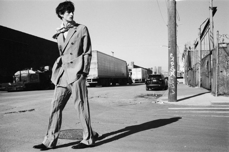 Monochrome image of a slender man in a suit crossing an urban street, with industrial buildings and a distant cityscape in the background.