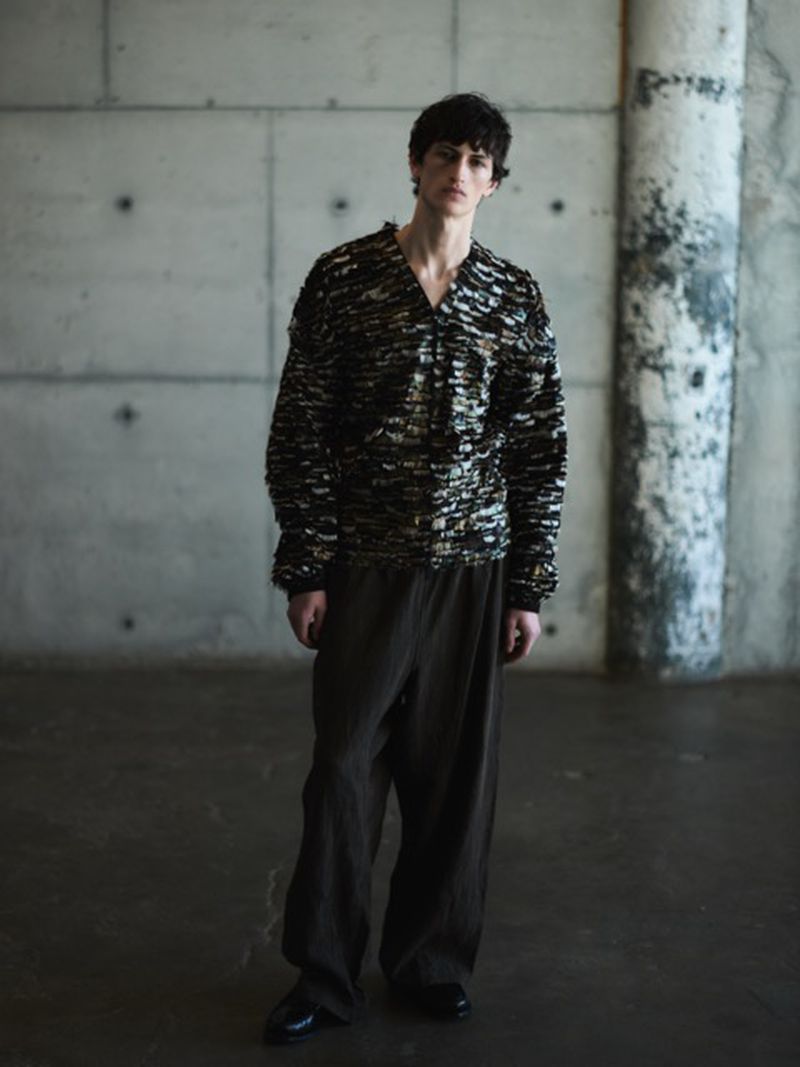 Male fashion model poses in a patterned sweater and loose-fitting pants against a textured grey concrete wall and floor backdrop.