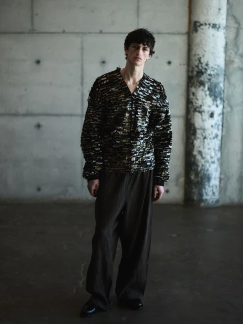 Male fashion model poses in a patterned sweater and loose-fitting pants against a textured grey concrete wall and floor backdrop.