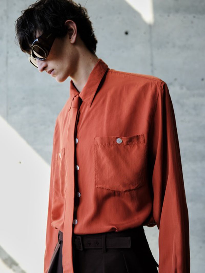 A brunette man wearing sunglasses poses indoors in a rust-colored button-down shirt and black pants against a concrete background.