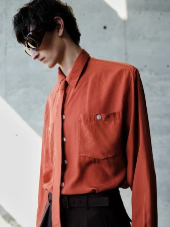 A brunette man wearing sunglasses poses indoors in a rust-colored button-down shirt and black pants against a concrete background.