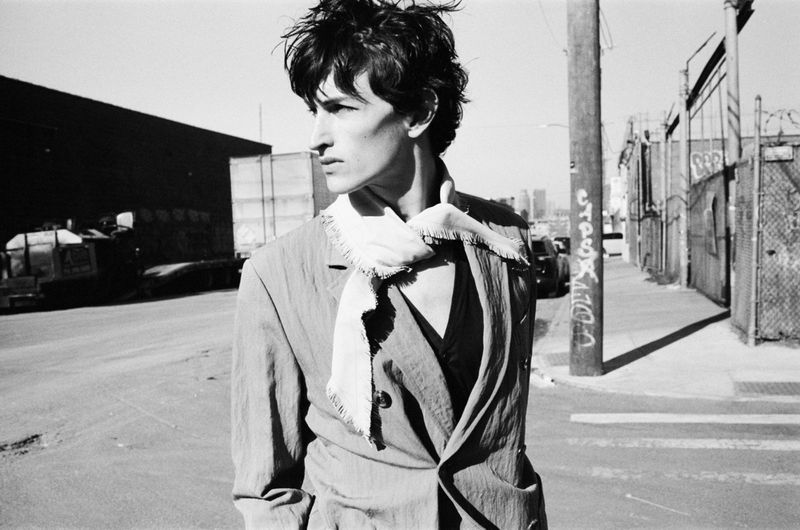 Monochrome portrait of an adult male in a suit standing on a street with industrial buildings, utility poles, and a city skyline in the distance.