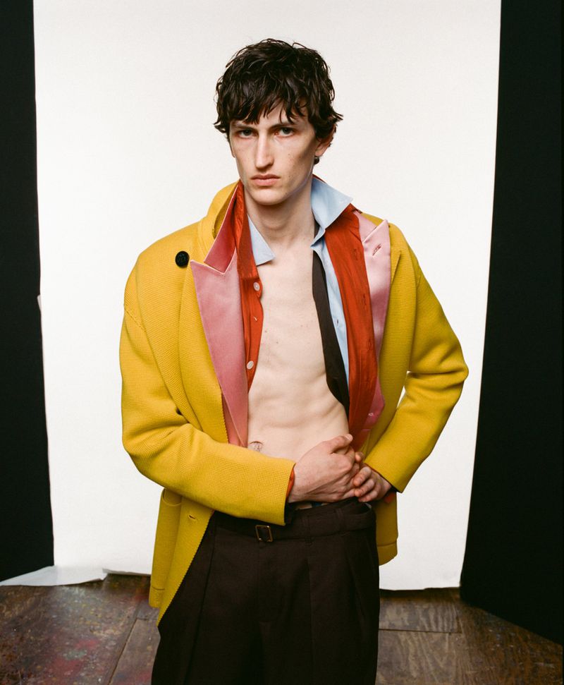 Male fashion model in a yellow overcoat, brown dress pants, and an unbuttoned shirt poses against a white photo studio backdrop.