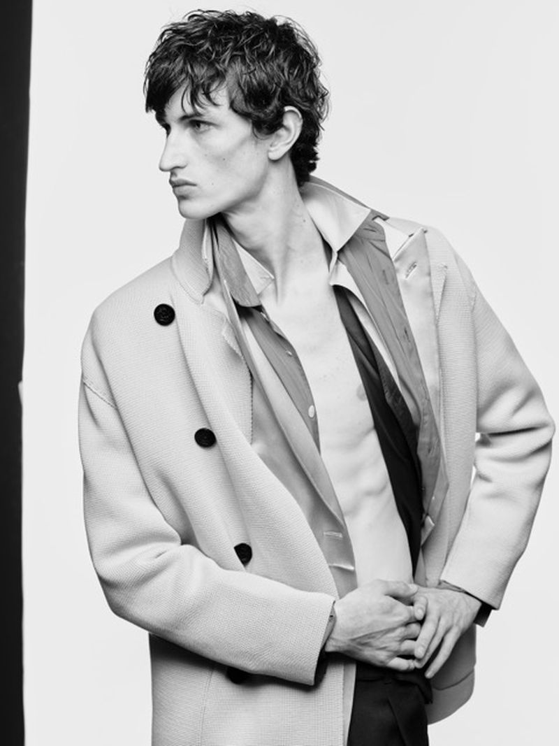 Monochrome portrait of a stylish male model with curly dark hair wearing a light-colored coat, shirt, and dark pants against a bright background.