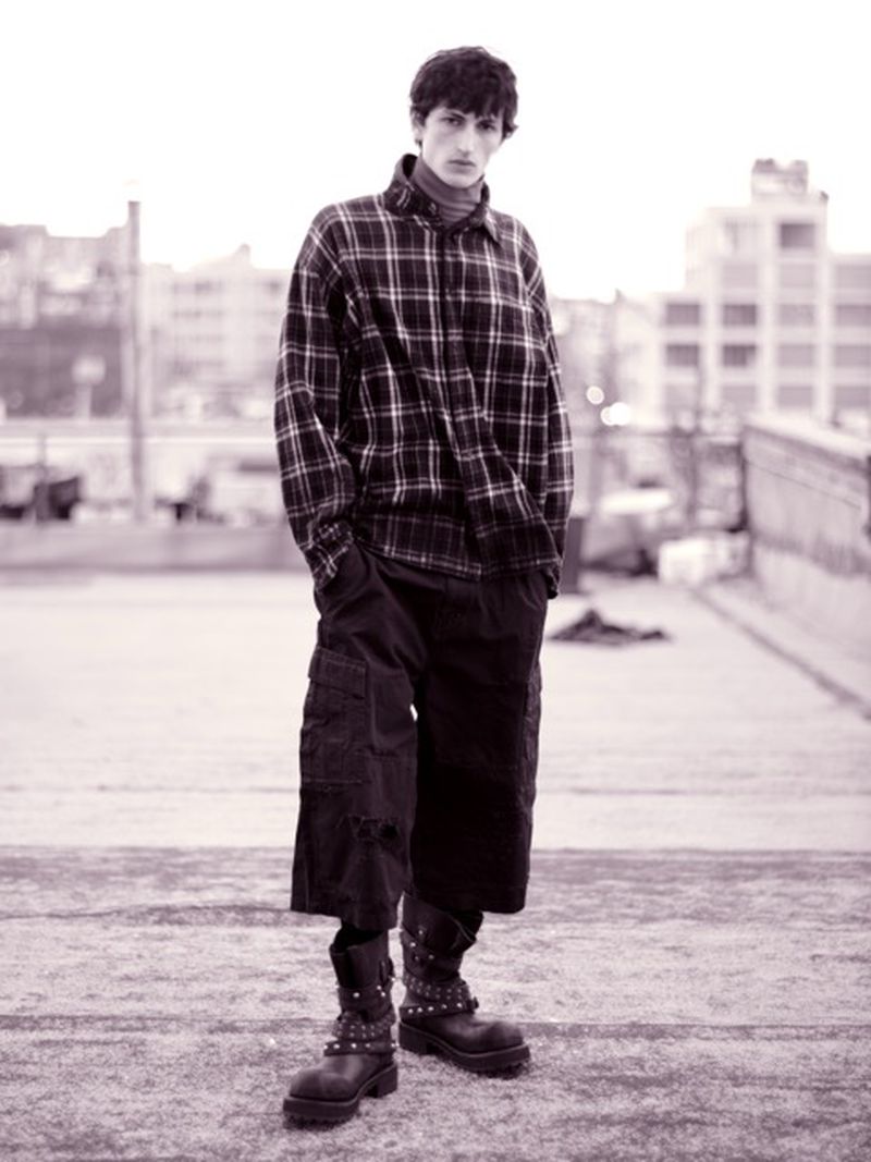 Monochrome portrait of a young adult man wearing a plaid shirt, oversized cargo pants, and studded boots, standing on a rooftop.