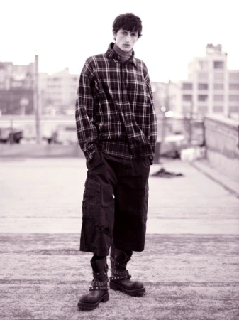 Monochrome portrait of a young adult man wearing a plaid shirt, oversized cargo pants, and studded boots, standing on a rooftop.