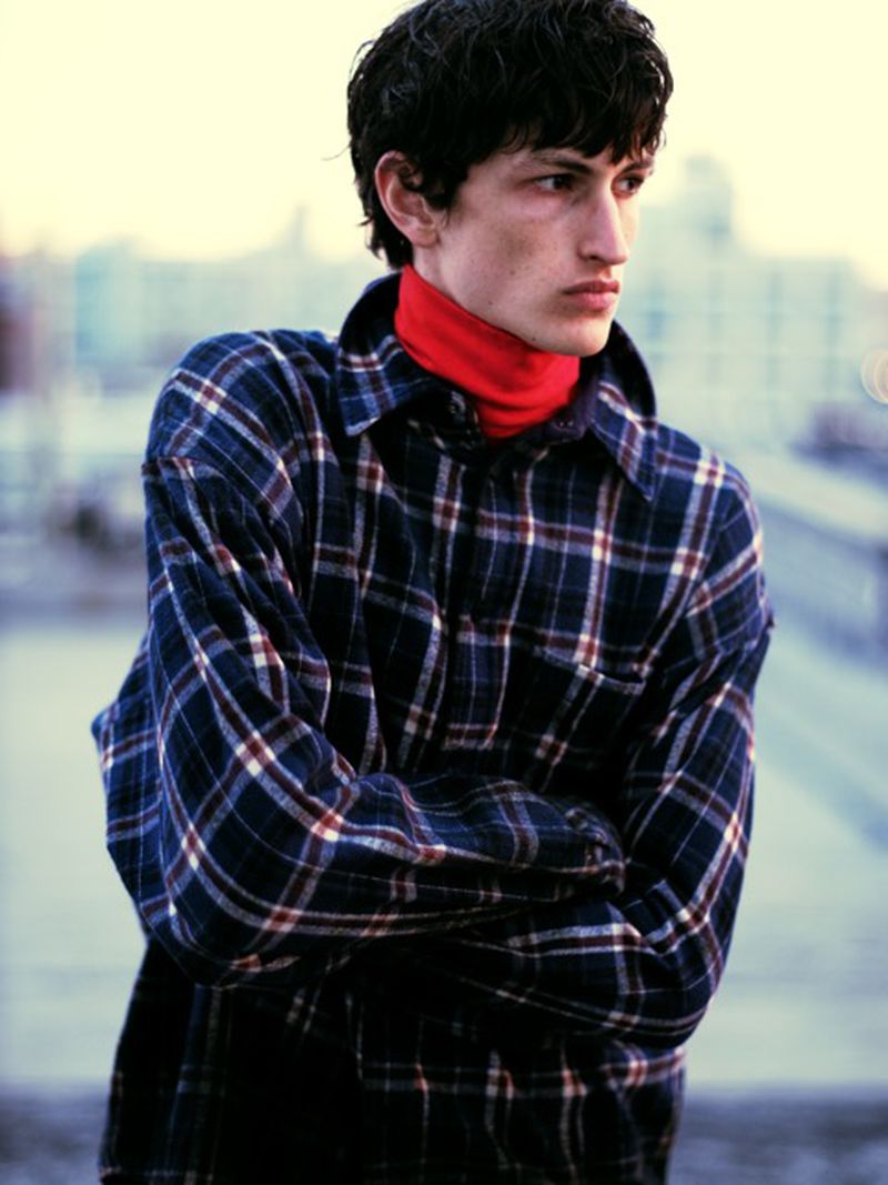 Young adult poses outdoors wearing a red turtleneck and a blue-and-white plaid jacket in a medium shot against a blurred cityscape.