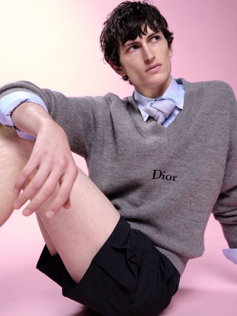 A young adult male poses in a photo studio, wearing a grey sweater with a Dior logo, a blue button-down shirt, tie, and black shorts against a pink backdrop.