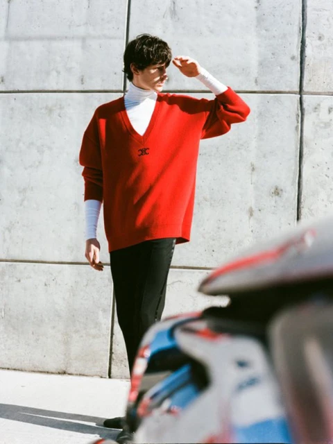 Male fashion model poses in red sweater and black pants against a concrete wall background, shielding his eyes from sunlight.