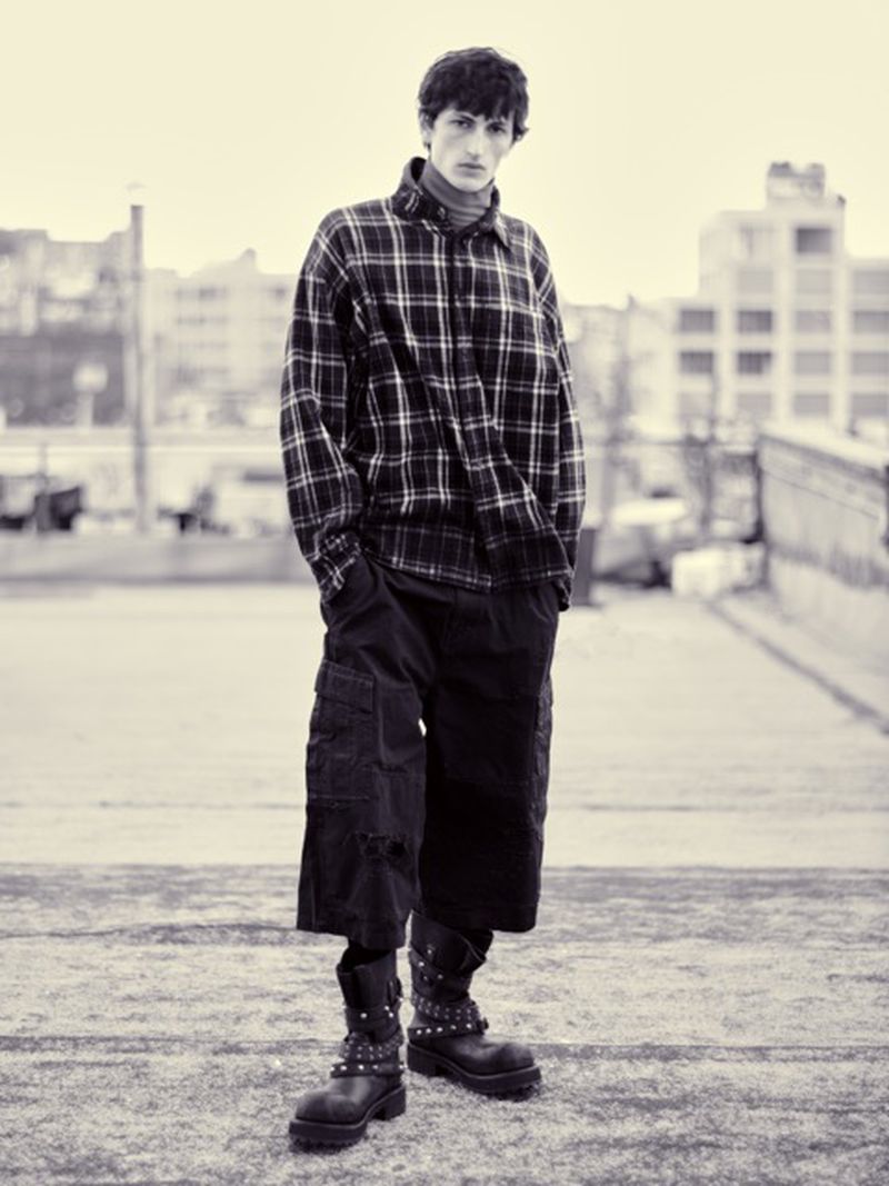 Monochrome portrait of a male fashion model in a plaid shirt, oversized pants, and studded boots posing on a rooftop with a cityscape background.