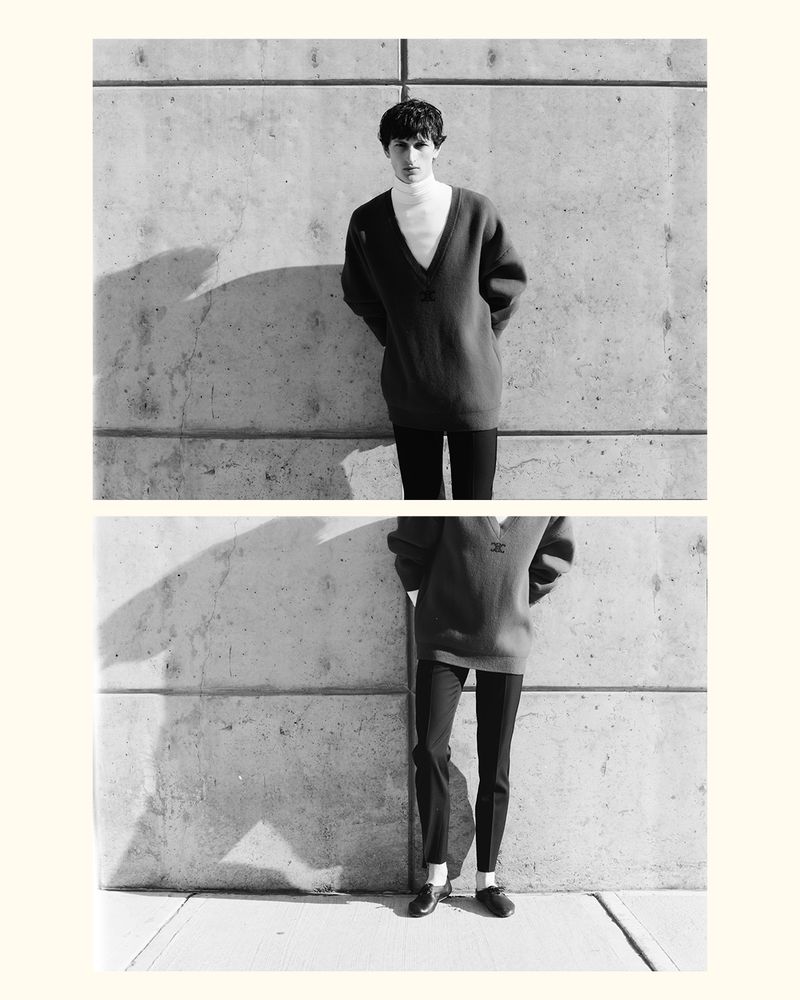 Monochrome shot of a young adult male in an oversized knit sweater and black pants posing against a concrete wall in an outdoor setting.