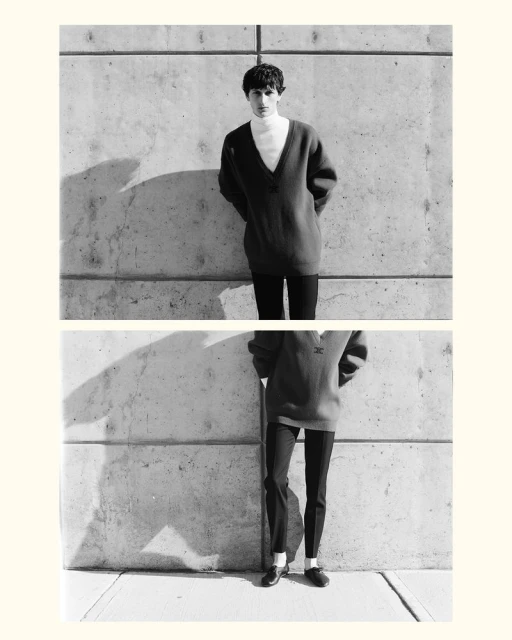 Monochrome shot of a young adult male in an oversized knit sweater and black pants posing against a concrete wall in an outdoor setting.