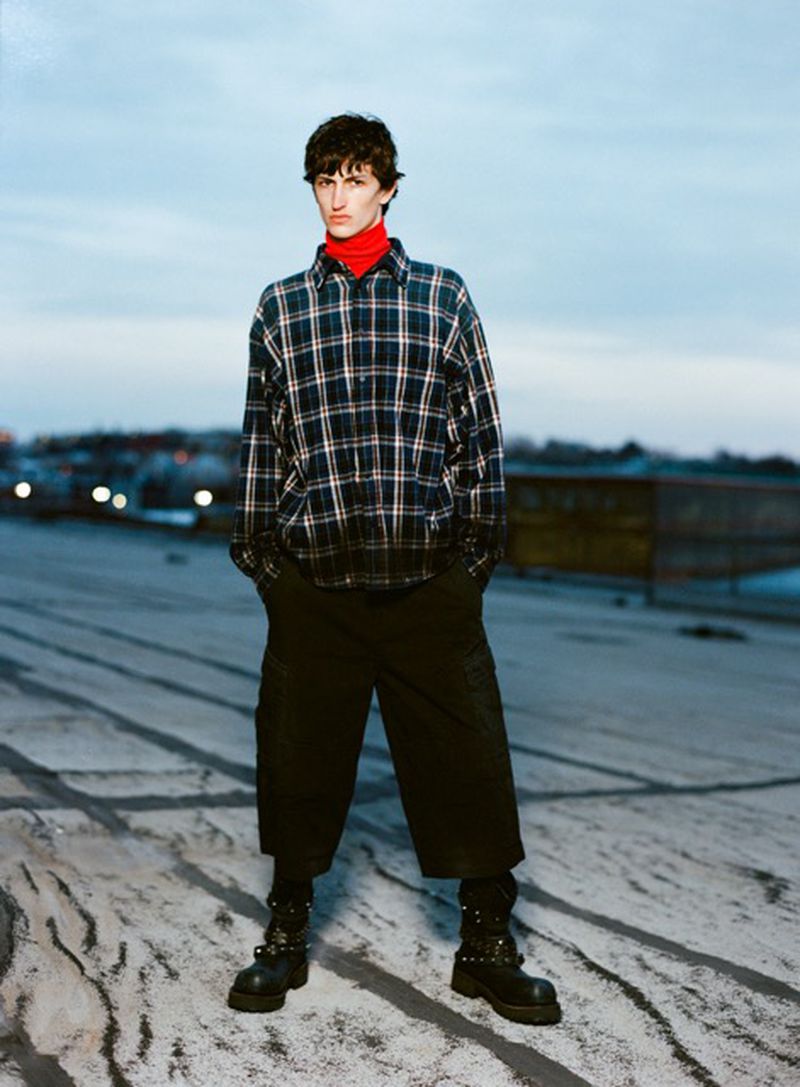 Young man in flannel and red turtleneck stands on a rooftop, wearing baggy pants and studded boots, against a light blue sky.