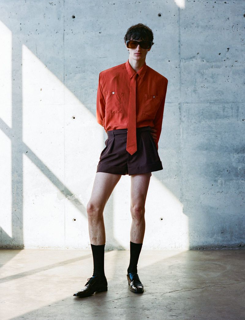 Fashion photo of an adult male model wearing a rust-colored shirt, brown shorts, black socks, and sunglasses standing indoors near a concrete wall.