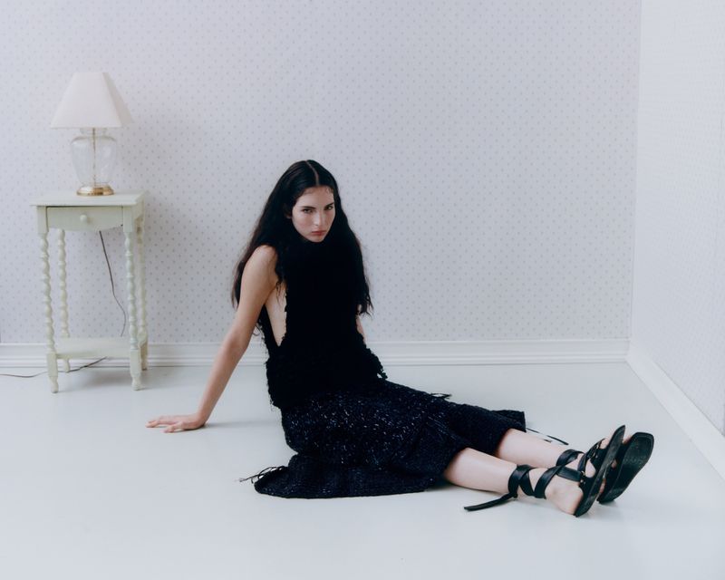 Woman with long dark hair poses on floor in black top, skirt, and strappy sandals next to lamp on table in front of patterned wall.