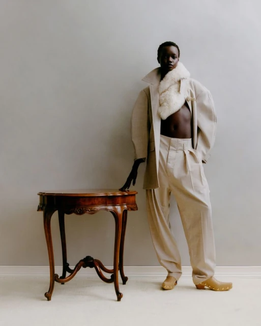 A striking editorial fashion portrait of a Black woman in a beige coat and pants, posed next to a vintage wooden table against a plain wall.