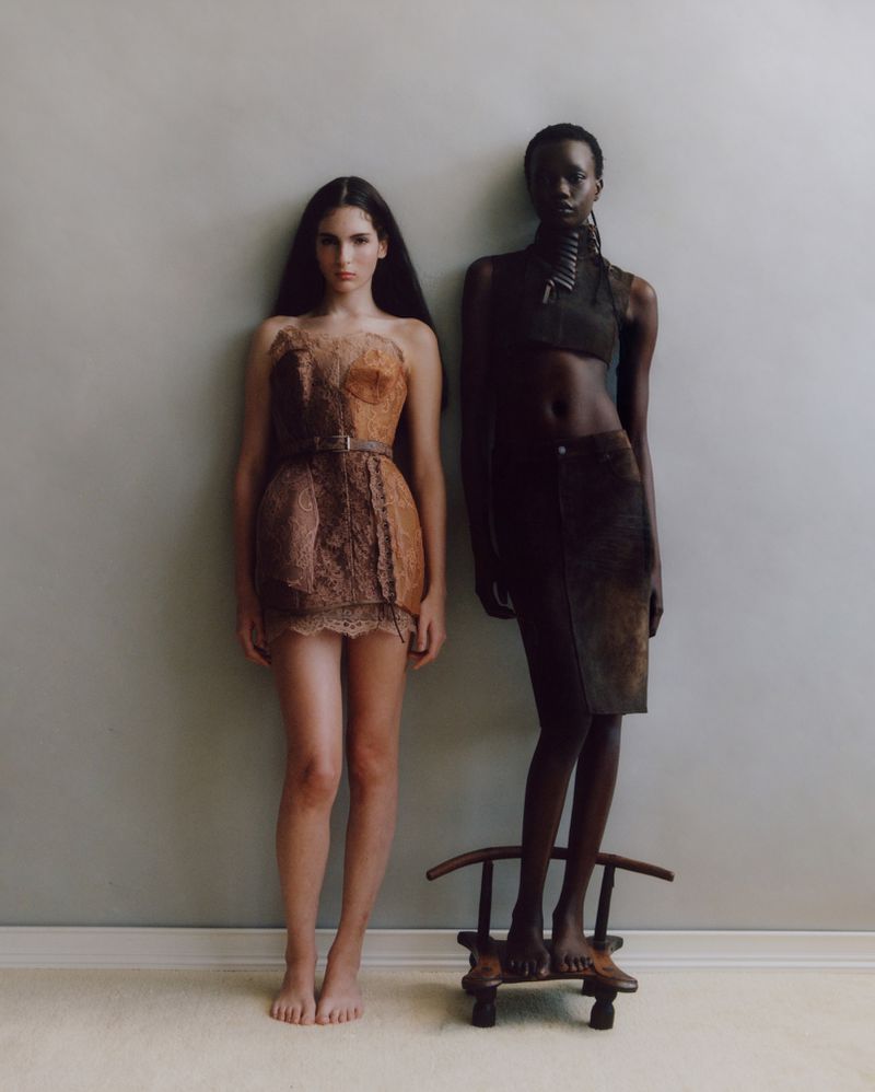 Two women in unconventional dresses pose in a studio, one standing on a small chair, against a neutral wall and light carpet.