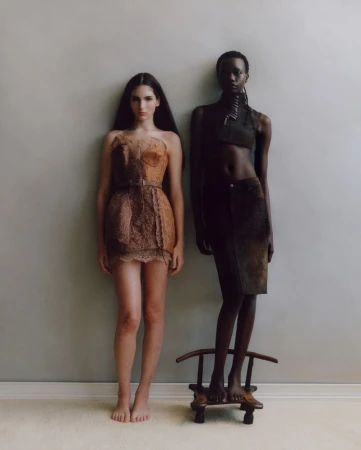 Two women in unconventional dresses pose in a studio, one standing on a small chair, against a neutral wall and light carpet.