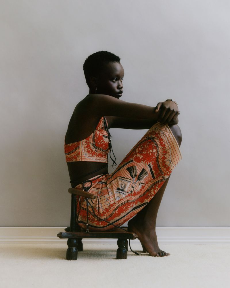 A young Black woman with short hair sits on a low stool in a patterned top and skirt, her arms resting on her knees, in a studio shot.