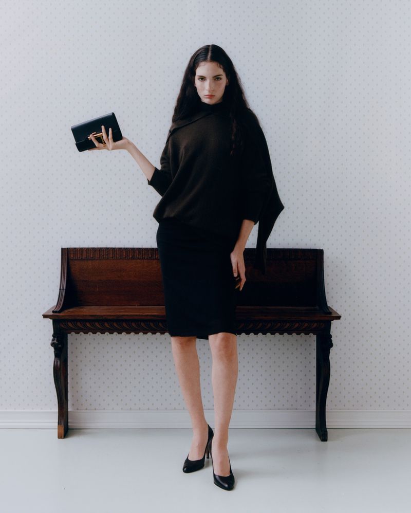 A woman in a dark sweater and skirt poses with a black clutch in a studio with a patterned wallpaper and a wooden bench.