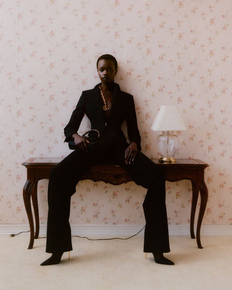 A fashionable Black woman in a black suit sits on a wooden console table with a glass lamp against a floral patterned wall.