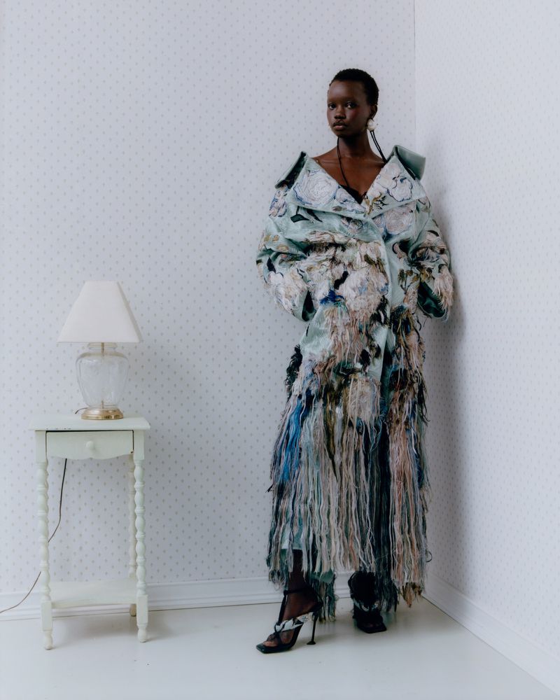 A Black female fashion model posing in an ornate, textured coat in a light-toned indoor corner setting with a bedside table and lamp.