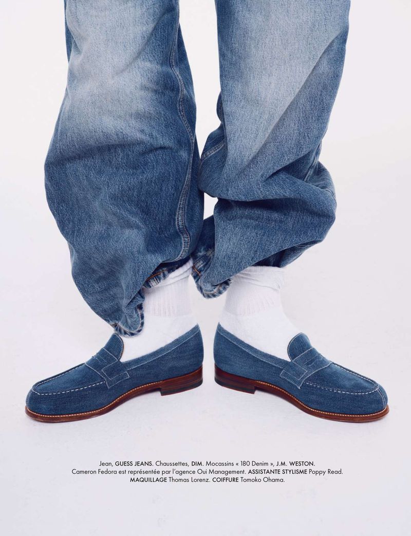 Close-up shot of blue suede loafers with white socks and cuffed denim jeans against a white backdrop in a studio fashion editorial.