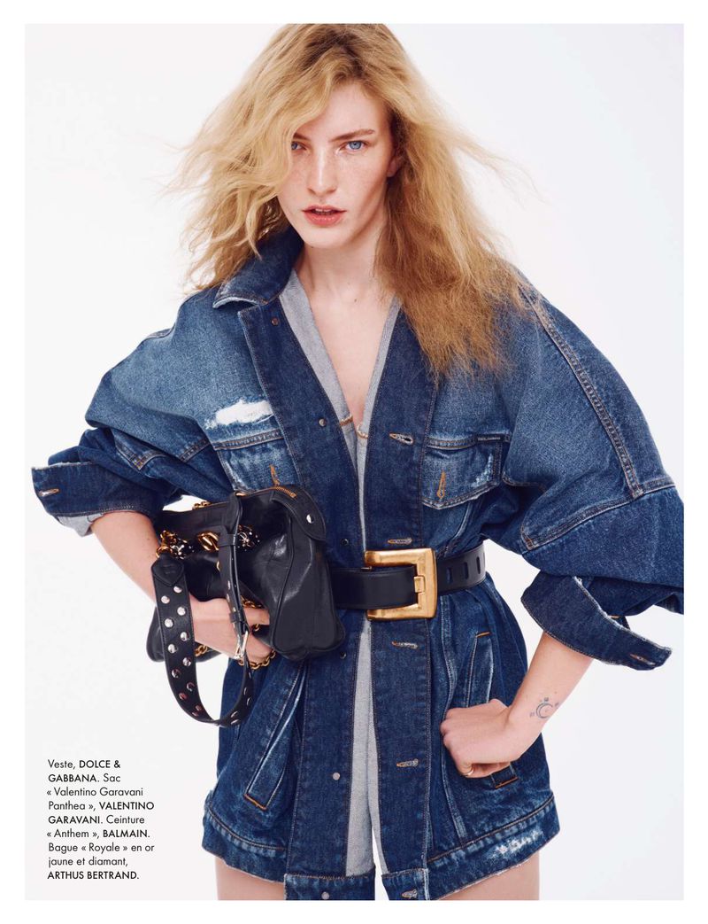 Fashion shot of a blonde adult woman wearing a denim jacket, large belt, and carrying a black studded bag against a white backdrop.