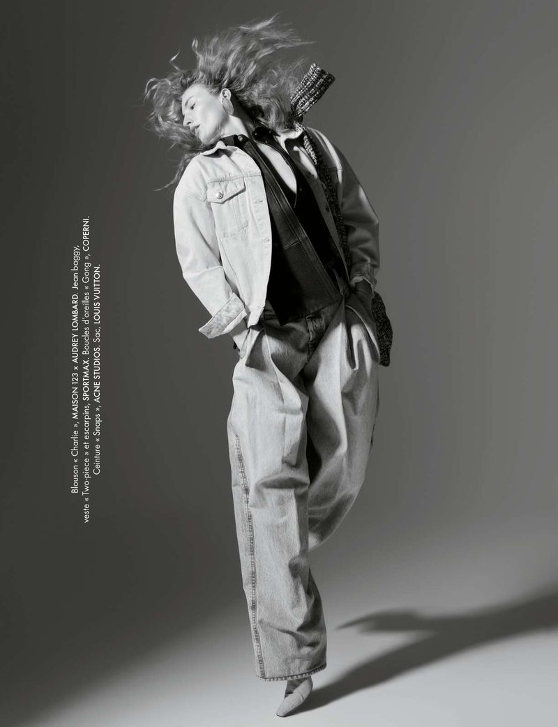 Monochrome studio shot of a stylish young woman in an unbuttoned denim jacket and wide-leg jeans, captured in mid-movement with flowing hair.