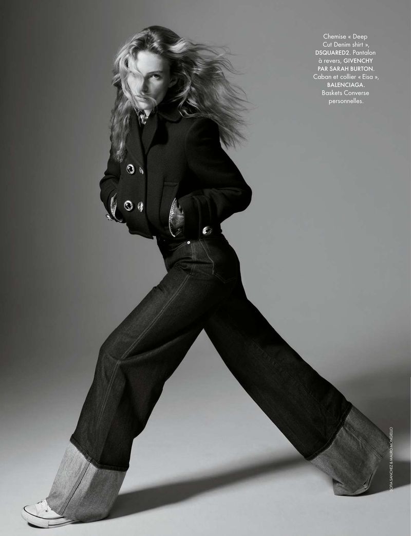 Editorial fashion shot of a young adult woman in a black jacket, denim pants and Converse sneakers, full-length pose against grey backdrop.