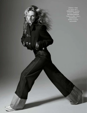 Editorial fashion shot of a young adult woman in a black jacket, denim pants and Converse sneakers, full-length pose against grey backdrop.