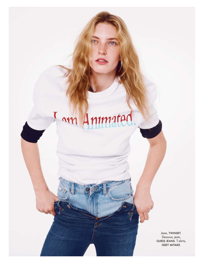 A fair-skinned young adult woman with blonde wavy hair poses in a studio wearing a t-shirt and unbuttoned blue jeans against a white backdrop.