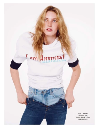 A fair-skinned young adult woman with blonde wavy hair poses in a studio wearing a t-shirt and unbuttoned blue jeans against a white backdrop.