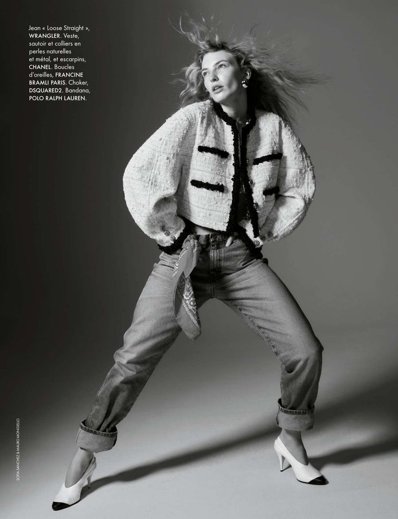 Adult woman in a studio wearing a tweed jacket, jeans, and heels. She has blonde hair and is posing in a dynamic stance.