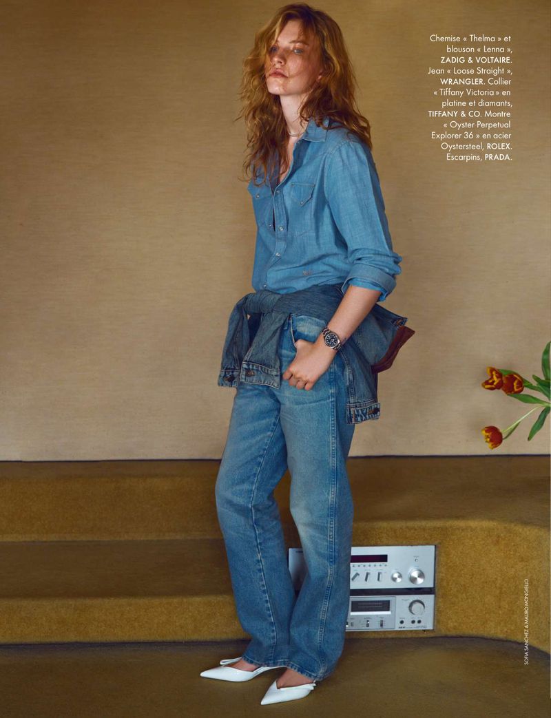 Full shot of a young adult woman with long hair wearing denim shirt and jeans, posed in front of neutral wall with amplifier.