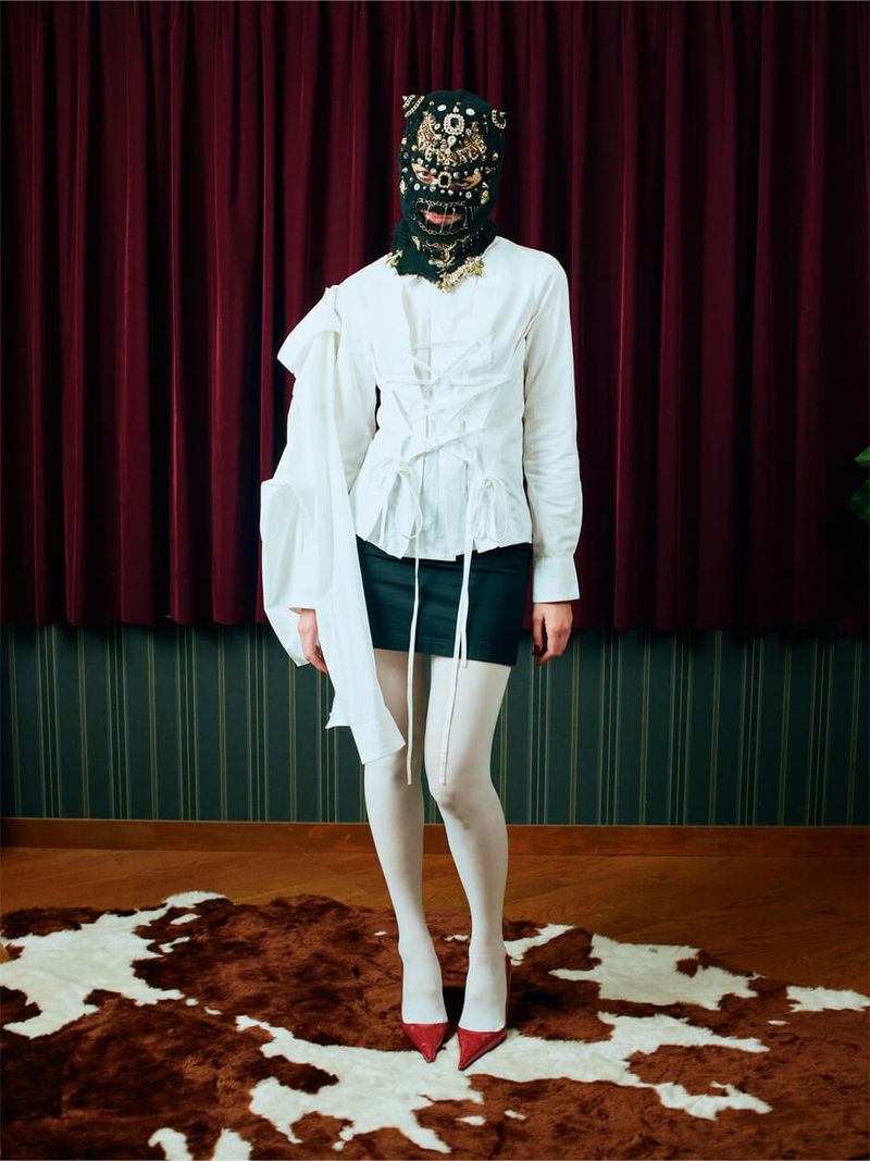A full-length portrait of a single adult female posing indoors wearing a white blouse, dark skirt, patterned mask, red heels, and white tights.