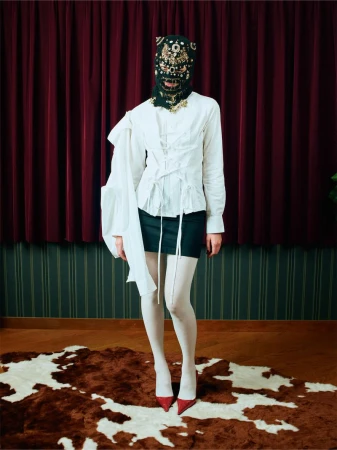 A full-length portrait of a single adult female posing indoors wearing a white blouse, dark skirt, patterned mask, red heels, and white tights.