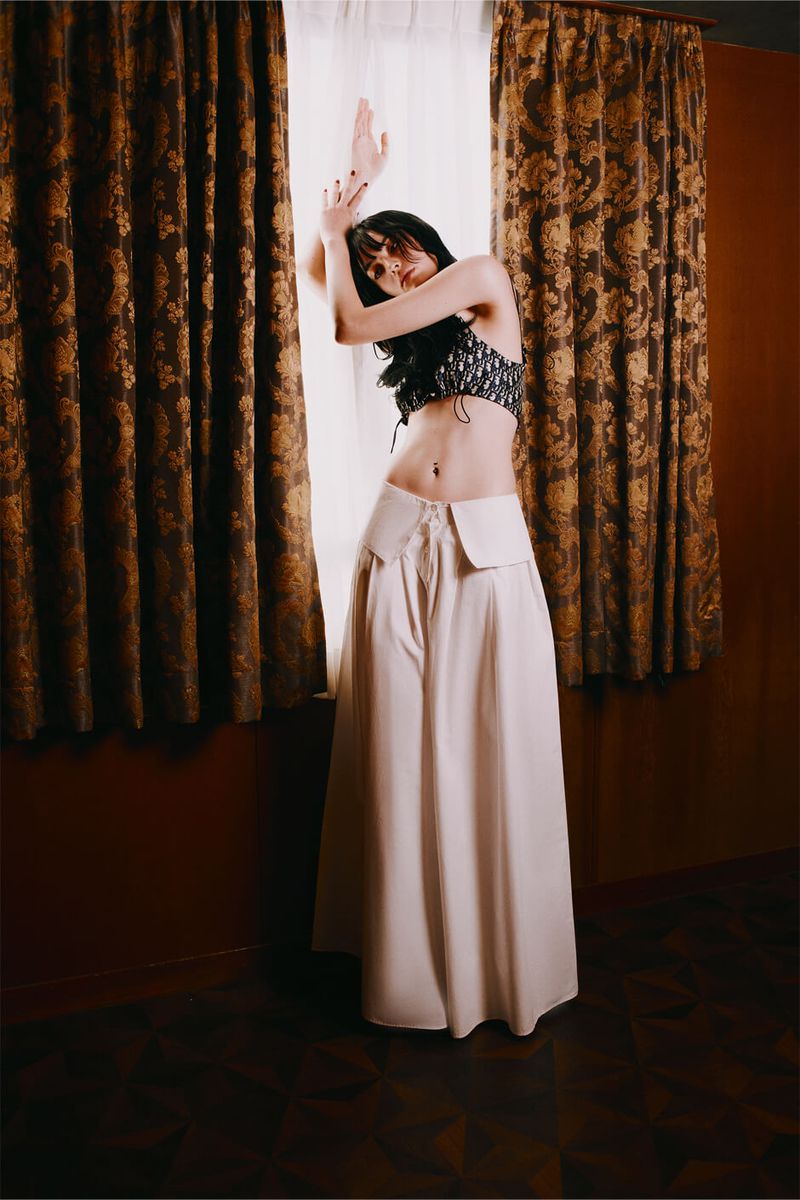 Young woman with dark hair poses indoors in a black top and long, light-colored skirt, leaning against a wall near a window and curtains.