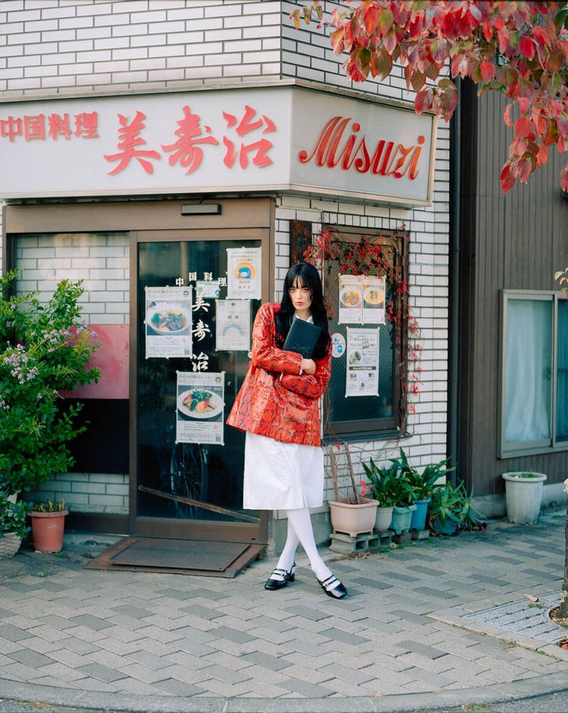 Adult woman in a red jacket and white dress stands in front of a Japanese restaurant with autumn foliage and brick sidewalk.