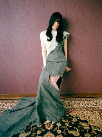 Elegant adult woman in a flowing grey, cream, and white asymmetrical dress posing against a pinkish-brown wall indoors on a patterned rug.