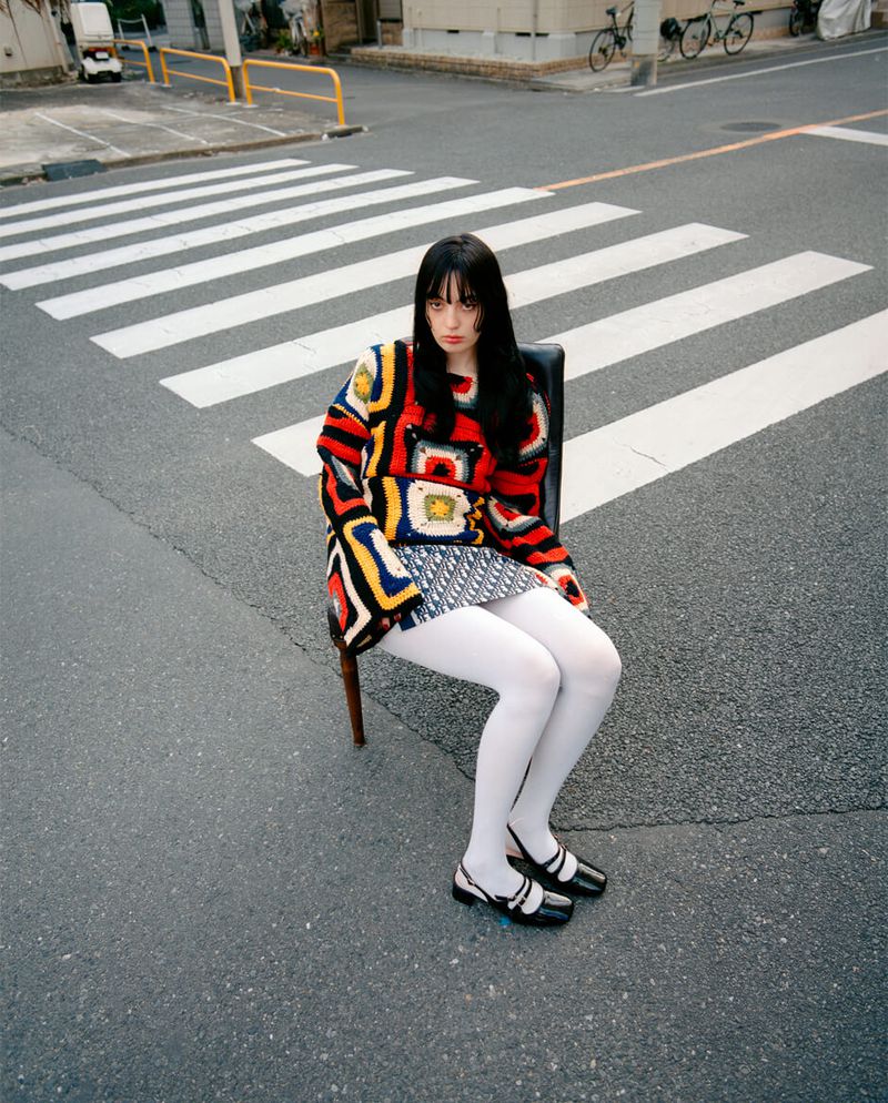 A young adult female with dark hair sits on a chair in the street with crosswalk, wearing a colorful knit sweater and black shoes.